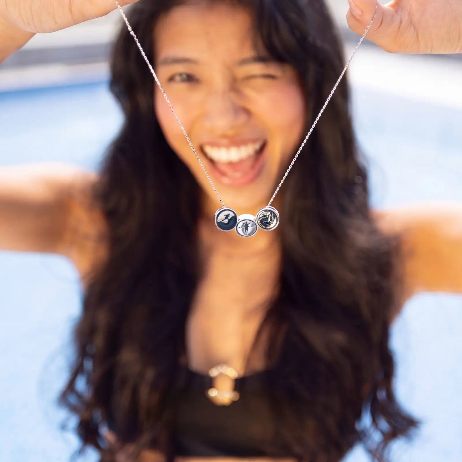 A smiling woman with long dark hair holds up a 925 sterling silver necklace with three circular Story Stroke charms in front of a blurred blue background, likely a poolside setting.